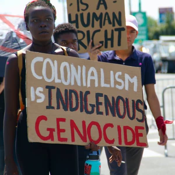 kultursensibel reisen: menschen mit demonstrationsschildern. auf einem steht: colonialism is indigenous genocide