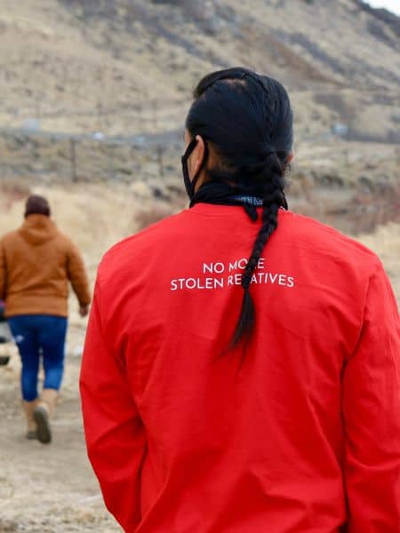 eine person mit langem schwarzem zopf und einer roten jacke von hinten, auf der steht "no more stolen relatives"